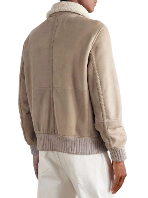 A back view of a man wearing a sophisticated tan suede bomber jacket, highlighting the plush cream shearling collar, detailed stitching across the back panel, and the ribbed knit hem and cuffs. The jacket is paired with light-colored trousers.