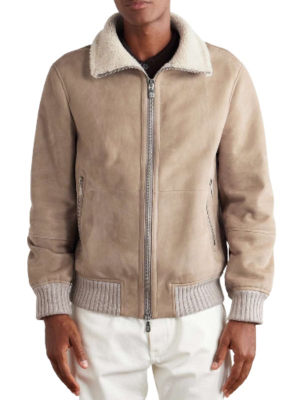 A stylish man, from the chest down, models a tan suede bomber jacket with a cream shearling collar and light grey ribbed knit details. He pairs the jacket with crisp white pants, presenting a refined and modern winter ensemble.