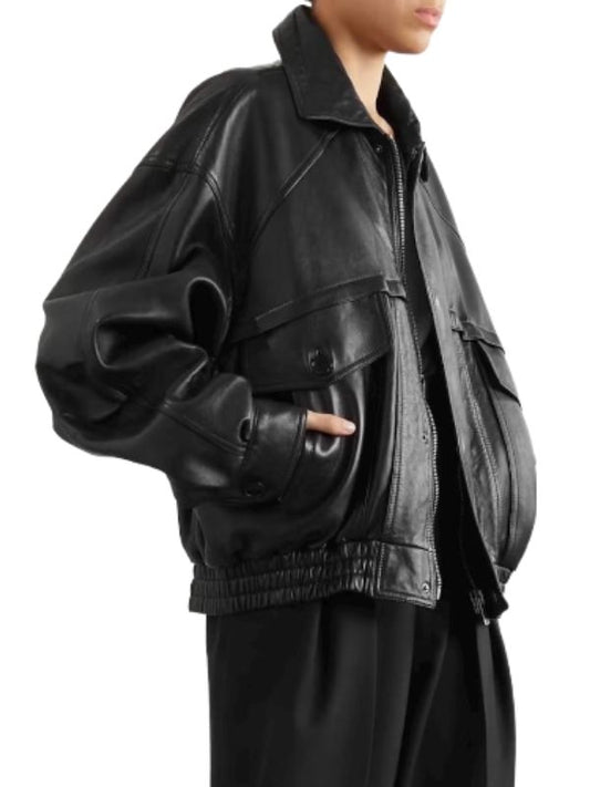 
A black, oversized, vintage-style leather bomber jacket is shown from the side. The jacket has a wide collar, a zippered front, and large, flap-covered pockets on the chest. The sleeves are long and gathered at the wrists, and the waistband is elasticated, creating a relaxed, voluminous silhouette. The leather appears soft and slightly distressed. The person wearing the jacket is also dressed in black, with a hint of their face visible at the top right.